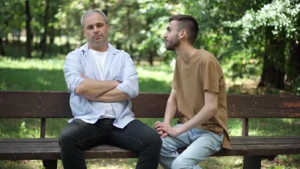 Alegre hijo adulto caucásico disculpándose como padre de mediana edad sentado en el banco con las manos cruzadas. Discutido familia en el soleado parque de verano al aire libre hablando. Conflicto y estilo de vida. — Vídeo de stock