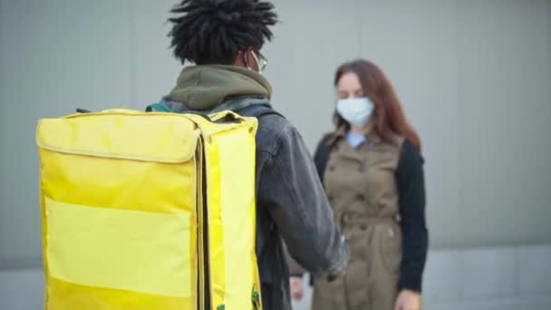 Rack focus va de courrier afro-américain masculin à la femme blanche dans Covid-19 masque de réception de colis à l'extérieur. Homme livrant la commande à un client en ville en cas de pandémie de coronavirus. — Video