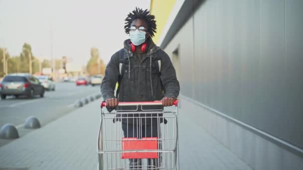 Portrét mužského kurýra s patami v Covidově masce tlačí nákupní vozík do obchodu. Afroameričtí mladí muži s taškou na jídlo pracující v městském městě. Koncept úlohy. — Stock video