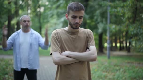 Joven culpable de pie en el parque de verano con las manos cruzadas como padres de mediana edad borrosa gritando y haciendo gestos en el fondo. Retrato de hijo ansioso molesto y padre regañando chico al aire libre. — Vídeos de Stock