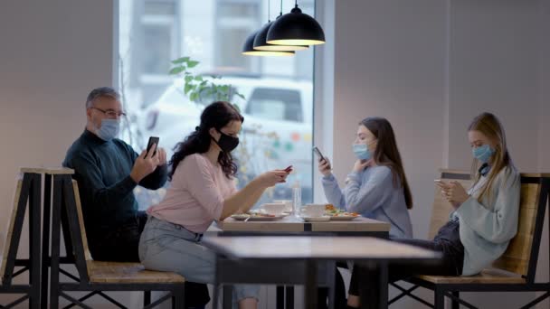 Generationenübergreifende Männer und Frauen in Covid-19-Gesichtsmasken surfen im Internet auf Smartphones im Café als verschwommene Kellnerin an der Front vorbei. Kaukasische Familie wartet im Restaurant auf Bestellung. — Stockvideo