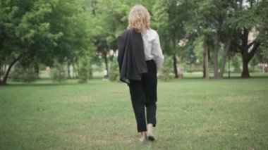 Kamera, parktaki yeşil yaz çimlerinde yürüyen sarışın beyaz iş kadınını takip ediyor. Kendine güvenen, gevşemiş, molada dışarıda gezinen bir kadının geniş açıyla görüntüsü. Yaşam tarzı ve özgüven.