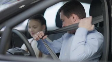 Kendine güvenen adam emniyet kemerini yavaş çekimde bağlıyor ve yolcu koltuğundaki bulanık tatlı kızla konuşuyor. Olumlu gülümseyen beyaz bir baba ve kızı araba galerisinde araç seçiyor. Otomobil endüstrisi.