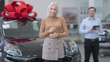 Çekici sarışın kadın kameraya dönüyor, arabanın anahtarlarını gösteriyor ve el ele tutuşuyor. Arka planda gülümseyen bulanık bir satıcı. Galeride poz veren mutlu, kendinden emin, yakışıklı, beyaz bir alıcının portresi..