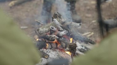 Ateş dışarıda ağır çekimde, erkek eli tahta sopayla yanıyor. Cephede ya da çokgende ateş etrafında oturan tanınmayan askerler. Savunma ve askeri kavram.