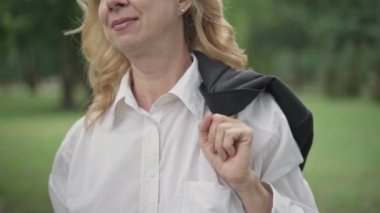 Tanımlanamayan orta yaşlı, ceketi omuzunda, yaz günü dışarıda yürüyen orta yaşlı bir kadının yakın çekimi. Pozitif gevşemiş Kafkas iş kadını parkta eğleniyor.