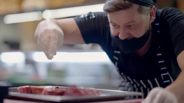 Zelfverzekerde professionele mannelijke kok in Covid gezicht masker zout rauw vlees op bakplaat in slow motion. Portret van chef-kok kokend smakelijk gerecht in restaurantkeuken. Man werkt in café aan pandemie. — Stockvideo