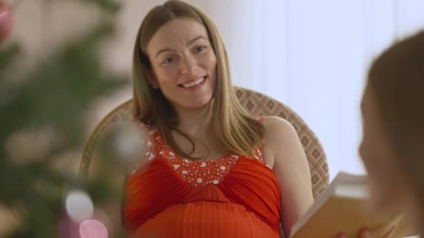 Красивая беременная брюнетка смеется и разговаривает с расплывчатой дочерью-подростком в канун Рождества в помещении. Портрет веселой расслабленной будущей кавказской матери, болтающей с девушкой на Новый год. — стоковое видео