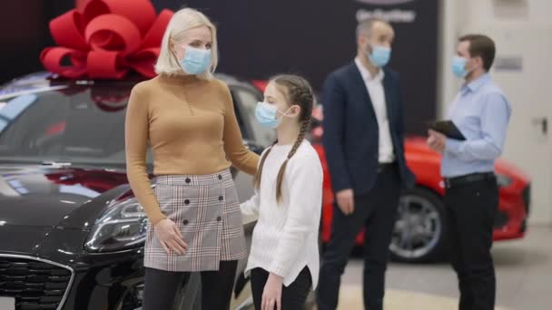 Covid-19 yüz maskeli mutlu beyaz anne ve kızı arka planda baba danışman satıcısıyla galeride poz veriyor. Varlıklı bir aile, galeride koronavirüs salgınıyla ilgili araç satın alıyor.. — Stok video
