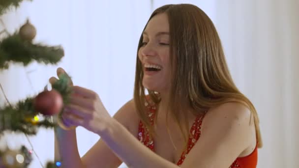 Mujer morena caucásica feliz decorando árbol de Año Nuevo en casa. Cámara se mueve de cara sonriente a vientre embarazada de joven alegre expectante disfrutando de la celebración de Navidad en el interior. — Vídeos de Stock