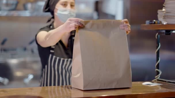 Unscharfe kaukasische junge Frau mit Covid-Gesichtsmaske, die in Cafés oder Restaurants Essen zum Mitnehmen einpackt. Professionelle Köchin bereitet bei Coronavirus-Pandemie in Zeitlupe Mittagstisch zu. — Stockvideo