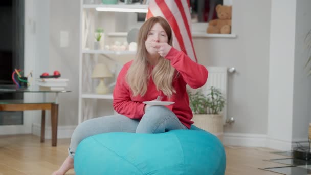 Entspanntes koreanisches Teenie-Mädchen vor dem Fernseher und beim Pizzaessen auf einem Stuhl im Innenraum. Weitwinkelporträt eines asiatischen Teenagers, der zu Hause die Freizeit genießt, mit amerikanischer Flagge im Hintergrund. Jugendalter. — Stockvideo