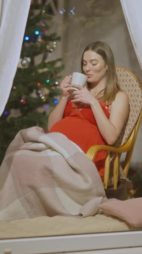 Video Vertikal, wanita hamil bahagia dengan gaun merah duduk di kursi goyang sambil minum teh panas dan melihat kamera di balik kaca jendela. Portrait of positive beautiful Caucasian expectant on New Year. — Stok Video