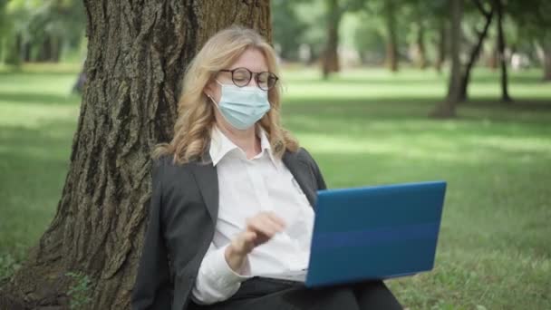 Foto del medio de alegre hermosa mujer caucásica en gafas y Covid-19 mascarilla portátil de cierre y mirando hacia otro lado sentado en el tronco del árbol en el parque de verano. Lady mensajería al aire libre sobre pandemia. — Vídeos de Stock