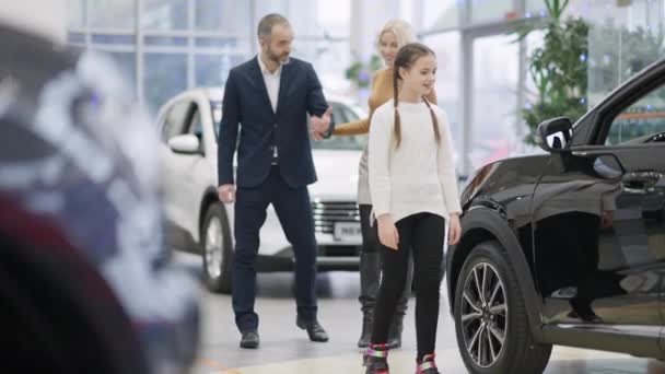 Széles kép egy lányról, aki autót választ egy kereskedésben pár szülővel. Portré boldog mosolygós gazdag kaukázusi lányról, apáról és anyáról, amint járművet vesznek a bemutatóteremben. Családi autóvásárlás. — Stock videók