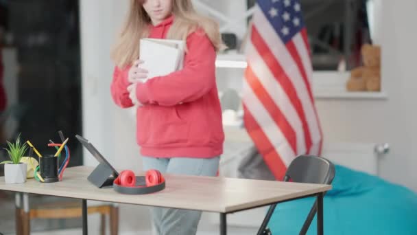 Zelfverzekerd Aziatisch meisje nadert tafel met boeken en gaat zitten. Portret van een gefocuste tiener die binnen studeert met Amerikaanse vlag op de achtergrond. Uitwisselingsstudent doet huiswerk. — Stockvideo