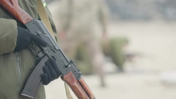 Detailní záběr armádní pistole v mužských rukou s rozmazanými vojáky střílejícími na pozadí na mnohoúhelník. Bělošští vojáci trénují venku na jaře nebo na podzim. Koncept zbraně. — Stock video
