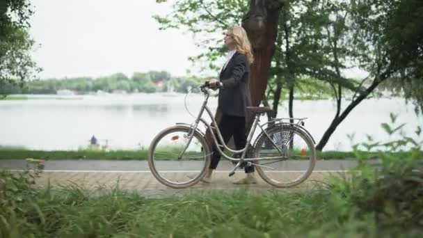 Πλευρική άποψη του ευτυχισμένη χαλαρή ευρωπαϊκή επιχειρηματίας περπάτημα με ποδήλατο κατά μήκος της ακτής λίμνη στο αστικό πάρκο. Ευρεία βολή της αυτοπεποίθηση λεπτή Καυκάσια ξανθιά μεσήλικη γυναίκα απολαμβάνοντας βόλτα σε εξωτερικούς χώρους. — Αρχείο Βίντεο