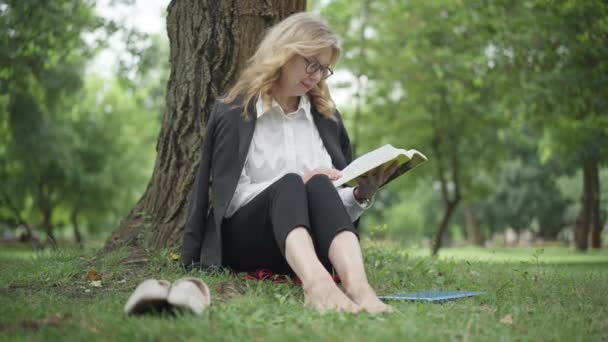 Amplio retrato de foto de mujer descalza de mediana edad feliz absorbida en anteojos sentados en el césped en el libro de lectura del parque de verano sonriendo. Positiva mujer de negocios caucásica disfrutando de hobby en el descanso al aire libre. — Vídeo de stock