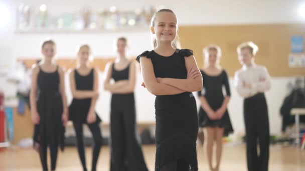 Portret uśmiechniętej dziewczyny pozującej ze skrzyżowanymi rękami w szkole tańca z rozmazanymi przyjaciółmi w tle. szczęśliwy kaukaski balet tancerz stojący w taniec studio patrząc w kamera. — Wideo stockowe
