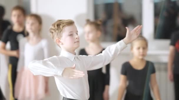 Portret pewnego siebie białego chłopca tańczącego walca w zwolnionym tempie mrugającego przed kamerą. Mała tancerka baletowa ćwicząca walca w spowolnieniu w szkole tańca z rozmazanymi przyjaciółmi w tle. Sztuka i umiejętności. — Wideo stockowe