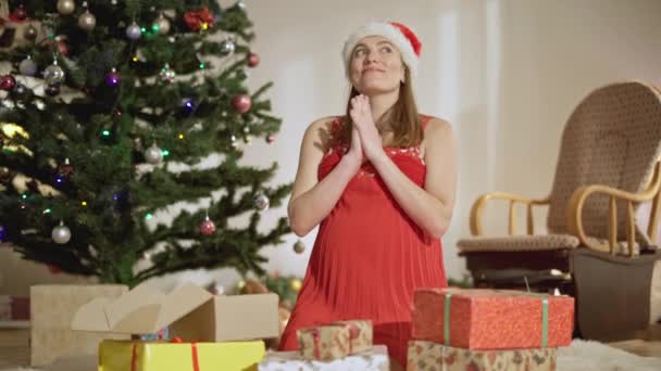 Joven mujer embarazada encantadora en sombrero de Navidad rezando por un mejor año sentado en casa con regalos alrededor. Retrato del expectante caucásico sonriente preparándose para las vacaciones. Esperanza y estilo de vida. — Vídeos de Stock