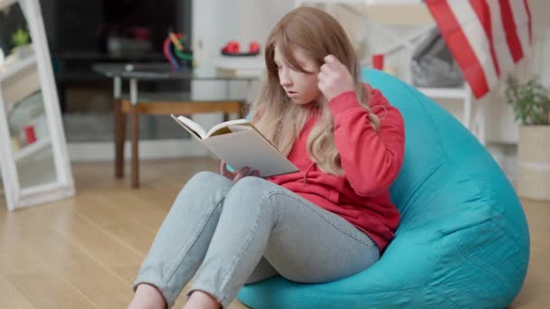 Concentré positif asiatique fille lecture à la maison et câlin livre. Portrait d'adolescent heureux insouciant appréciant la littérature à l'intérieur. Redhead immigrant reposant avec drapeau des États-Unis à l'arrière-plan. — Video