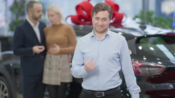 Dealer auto caucazian mulțumit care arată da gest în mișcare lentă și zâmbind uitându-se la camera foto cu cuplu neclar cu automobil cadou pe fundal. Portretul vânzătorului profesionist în dealer . — Videoclip de stoc
