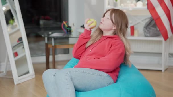 Relajada adolescente asiática comiendo manzana saludable sentada en la silla de la bolsa en casa. Retrato de adolescente lindo mordiendo y masticando fruta en el interior. Estilo de vida saludable y adolescencia. — Vídeos de Stock