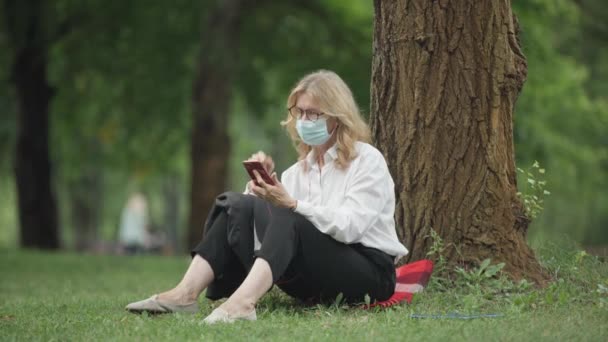 Wide shot of relaxed middle aged Caucasian woman in Covid-19 face mask sitting at tree in park listening to music from smartphone playlist. Blond businesswoman resting outdoors on coronavirus pandemic — Stock Video