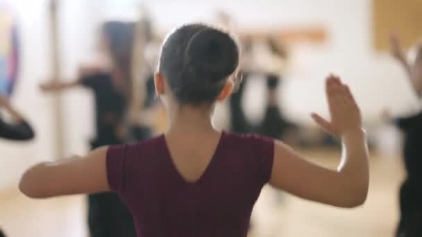Bruneta běloška s hnědýma očima poskakuje a točí se na čtvrté pozici v taneční škole. Rozmazané děti nacvičují v pozadí. Lekce v tanečním studiu pro děti uvnitř. — Stock video