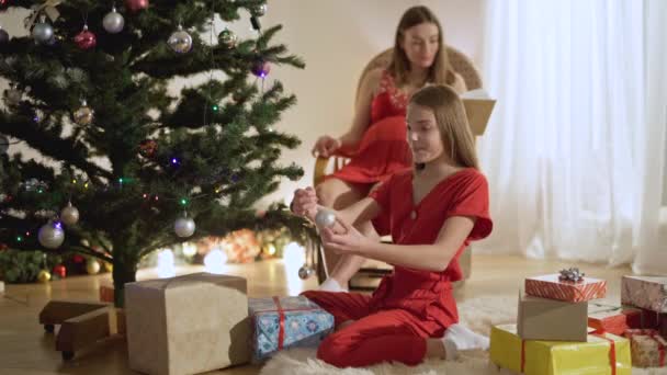 Bela adolescente decorando árvore de Ano Novo com mãe grávida lendo livro no fundo. Feliz filha caucasiana ajudando a mãe na véspera de Natal em casa e beijando o pai. Conceito familiar. — Vídeo de Stock