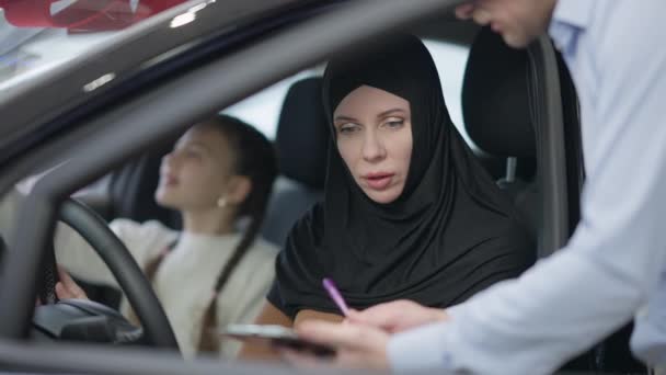 Mujer hermosa de confianza en hijab firma acuerdo de compra y venta sentado en el asiento del conductor en la sala de exposición. Retrato de una señora rica y hermosa comprando un automóvil en concesionario con su hija. — Vídeo de stock