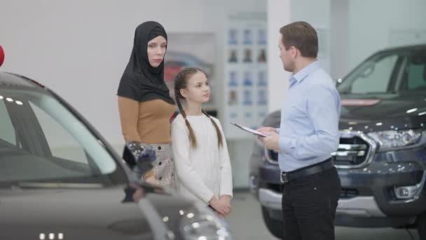 Boční pohled na auto dealer poradenství žena v hidžábu a hezká dívka v showroomu. Profesionální zaměstnanec pomáhá s výběrem nového automobilu v autosalonu mluvení a gestikulace. Zpomalený pohyb. — Stock video