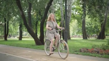 Neşeli, sarışın, orta yaşlı bir kadının güneşli yaz parkında bisiklete binip durduğu geniş bir kare. Dışarıda bisiklete binmekten hoşlanan heyecanlı beyaz kadın portresi. Hobi ve sağlıklı yaşam tarzı.
