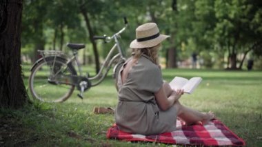 Dertsiz, beyaz, sarışın, orta yaşlı, kitap okuyan bir kadının park alanındaki yaz otlağında oturması. Olumlu, rahat okuyucu dışarıda hobi ediniyor. Keyif ve yaşam tarzı.