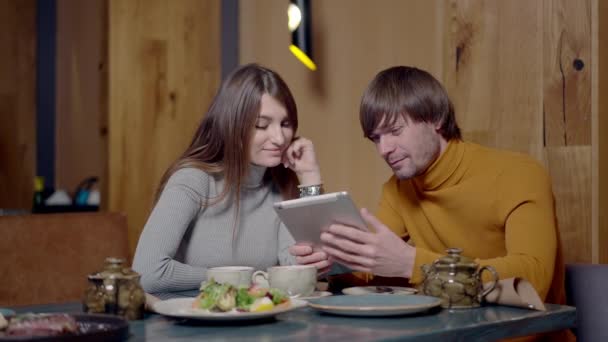 Középső felvétel portré boldog mosolygós fiatal kaukázusi pár szörfözés internet tabletta és beszél. Jóképű Y-generációs férfi és gyönyörű nő randizik az étteremben. Lassú mozgás.. — Stock videók