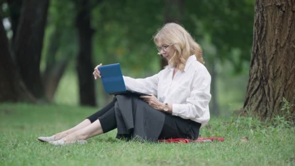 Widok z boku pewnej siebie bizneswoman w okularach otwarcia laptopa siedzi na zielonej łące na zewnątrz i wpisując na klawiaturze. Szerokie ujęcie eleganckiej szczupłej kobiety w średnim wieku wiadomości online w parku. — Wideo stockowe