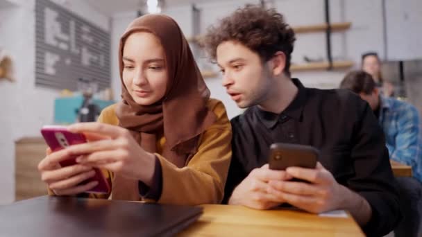 快乐的一对年轻的穆斯林夫妇坐在咖啡馆里，用智能手机上的社交媒体信息。笑着英俊的男人和漂亮的女人在网上冲浪，在自助餐厅聊天。千年生活方式. — 图库视频影像