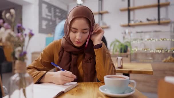 Koncentrált fiatal muszlim nő telefonál és ír egy kávézóban elmosódott kaukázusi pincérnővel Covid maszkban, aki ügyfelet szolgál ki a háttérben. Zseniális diák vagy üzletasszony portréja. — Stock videók