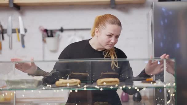 Retrato de media toma del trabajador positivo de la cafetería tomando postre dulce en el mostrador y alejándose. Mujer caucásica de talla grande que trabaja en la cafetería. Vendedor en el lugar de trabajo interior. — Vídeo de stock