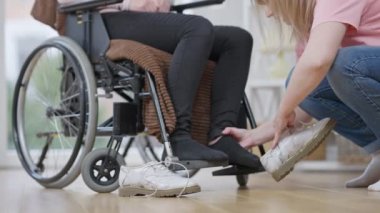 Kafkasyalı genç bir kadının tekerlekli sandalyede oturan engelli birinin ayaklarına ayakkabı giymesi. Gönüllü ya da arkadaşı belden aşağısı felçli bir kadına yardım ediyor. Bakım ve sakatlık kavramı.