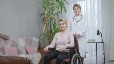 Neşeli beyaz doktor portresi ve evde poz veren özürlü hasta. Pozitif güzel doktor ve tekerlekli sandalyedeki felçli kadın gülümseyen kameraya bakıyor. Yaşam tarzı ve ilaç.