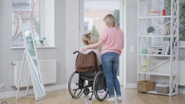 Tekerlekli sandalyedeki pozitif engelli kadının arkadaşlarıyla gülümsediği geniş bir bakış açısı. Mutlu Kafkasyalı felçli, sakat, evde genç bir bayanla dedikodu yapıyor. Dostluk ve hastalık.