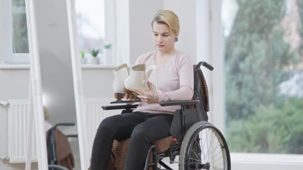 Jeune femme caucasienne frustrée en fauteuil roulant regardant les patins à glace et la pensée. Portrait d'une sportive paralysée handicapée aux jambes blessées. Invalidité et paralysie. — Video