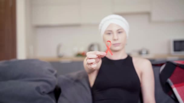 Närbild av smal kvinnlig hand håller rött band anti-cancer symbol med suddig allvarlig ung kvinna i bakgrunden tittar på kameran. En vacker dam som slåss med sjukdomar. Onkologi och hälsovård. — Stockvideo