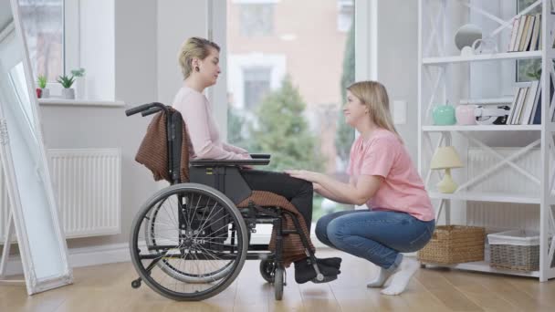 Vista laterale ampio scatto di giovane donna disabile paralizzata sulla sedia a rotelle parlare con amico solidale a casa. Sconvolto paraplegico caucasico non valido supportato da un amico in casa. Stile di vita della disabilità. — Video Stock