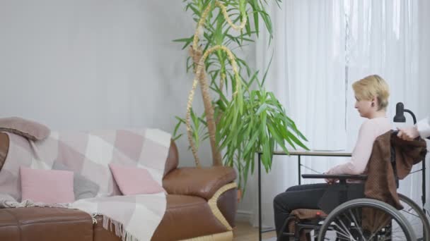 Portrait large de l'infirmière ou du bénévole roulant en fauteuil roulant avec une femme handicapée dans la chambre et assise sur le canapé. Aide soignante prenant soin d'un patient caucasien paralysé paraplégique à l'intérieur. Mouvement lent. — Video