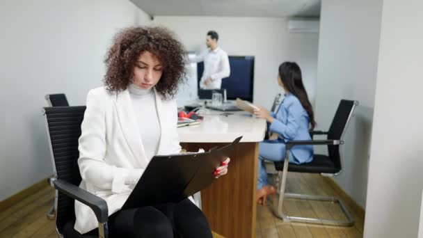 Jonge, geconcentreerde blanke vrouw die documenten analyseert die aan vergadertafel zitten met multiraciale collega 's die op de achtergrond praten. Professionele werknemer die binnen werkt. — Stockvideo