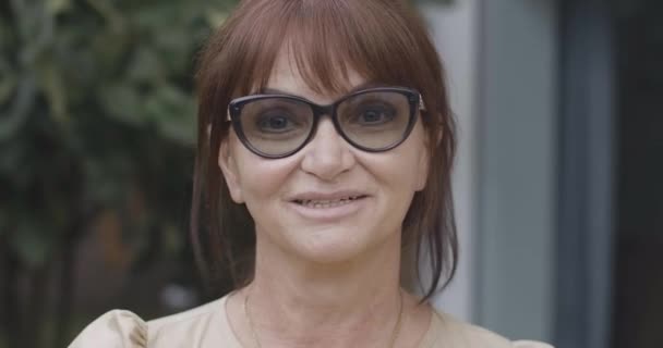 Captura de cabeça de mulher sênior confiante bonita em óculos olhando para a câmera e sorrindo. Close-up retrato de ruiva encantador aposentado com olhos cinzentos posando ao ar livre no dia de verão. Cinema 4k ProResHQ — Vídeo de Stock